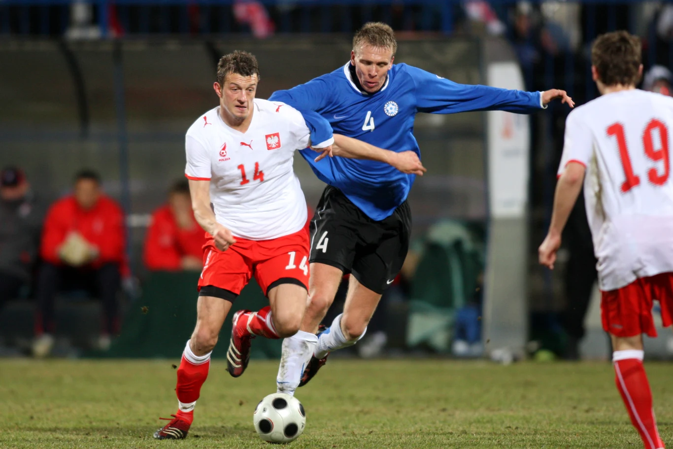 zawodnik w białoczerwonym stroju z numerem 14 prowadzi piłkę, naciskany przez przeciwnika w niebieskiej koszulce z numerem 4, obok widoczny kolejny piłkarz w czerwonych spodenkach i białej koszulce z numerem 19