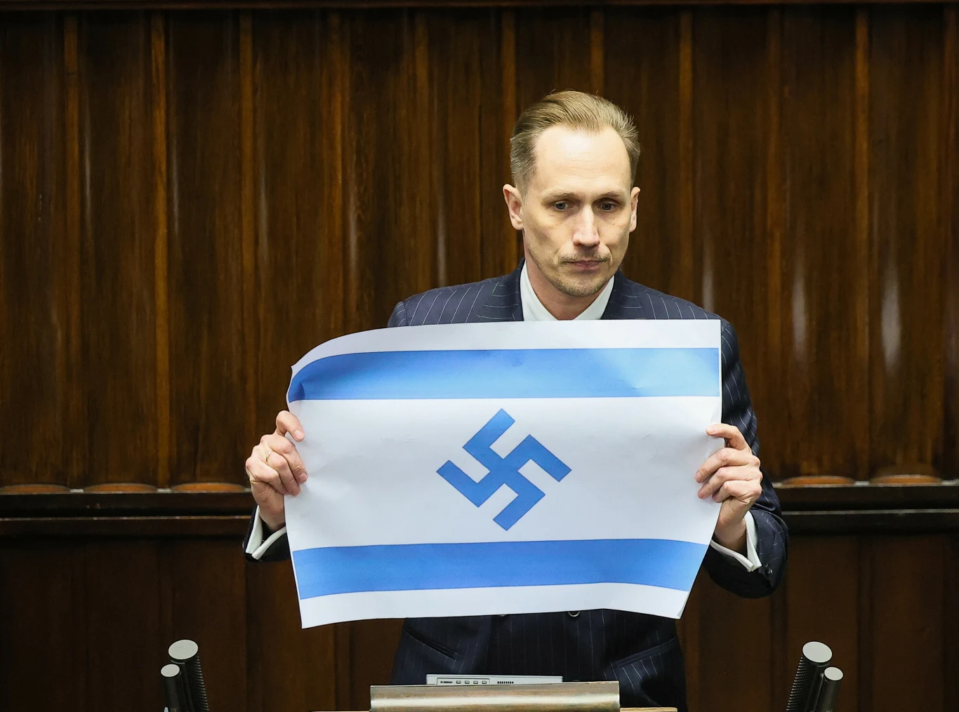 Konrad Berkowicz w Sejmie Mężczyzna w garniturze stoi za mównicą, trzymając plakat z niebiesko-białą flagą, na której centralnie umieszczona jest niebieska swastyka.