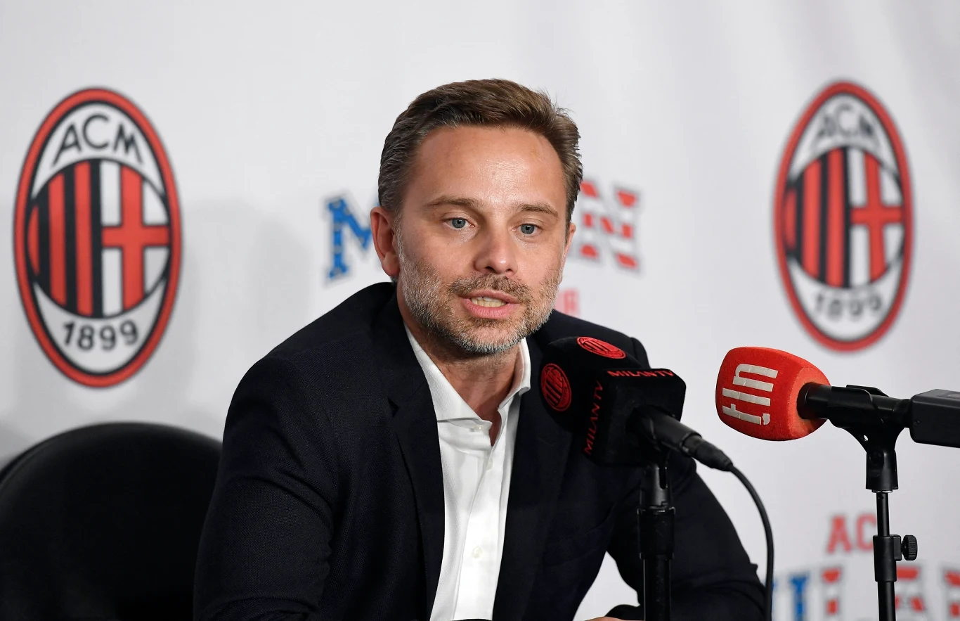 mężczyzna w garniturze siedzący przy stole konferencyjnym, przemawiający do kilku mikrofonów, w tle widoczne logo AC Milan i inne logotypy na ściance prasowej