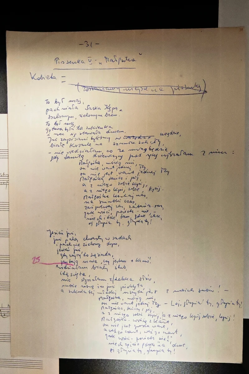 Rękopis tekstu piosenki napisany niebieskim długopisem na pożółkłej kartce papieru, zawierający poprawki i przekreślenia, a w lewym dolnym rogu widoczny fragment nutowego arkusza.