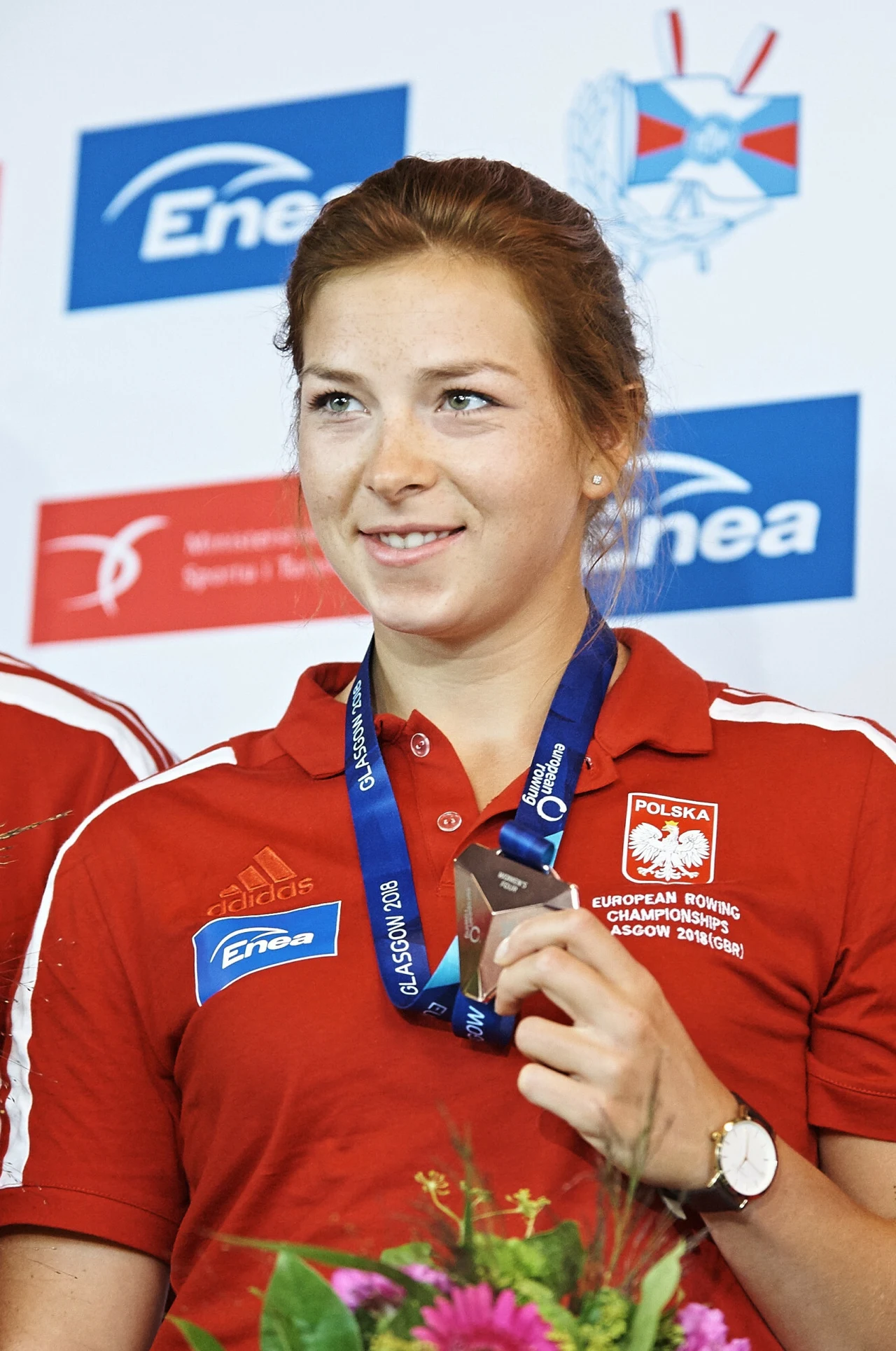 Olga Michałkiewicz kobieta w czerwonej koszulce reprezentacji Polski z logo Enea, trzymająca medal na niebieskiej wstążce, uśmiechnięta, z bukietem kwiatów na pierwszym planie i tłem z logotypami partnerów