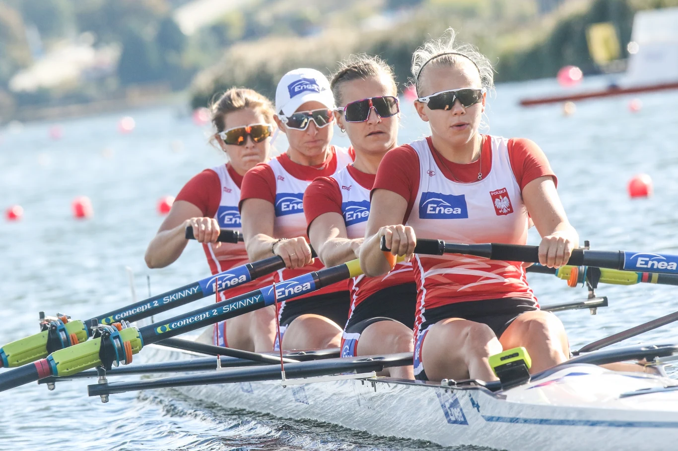 Maria Wierzbowska, Olga Michałkiewicz, Monika Chabel, Paulina Grzella Cztery kobiety w czerwono-białych strojach sportowych wiosłują synchronicznie w łodzi wioślarskiej podczas zawodów na wodzie, skupione i skoncentrowane na wysiłku, z widocznymi logotypami partnerów na wiosłach i koszulkach.
