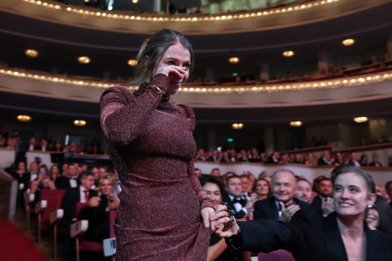 Klaudia Zwolińska Klaudia Zwolińska w cekinowej sukni przechodzi przez teatr, trzymając osobę za rękę, wśród klaszczącej publiczności