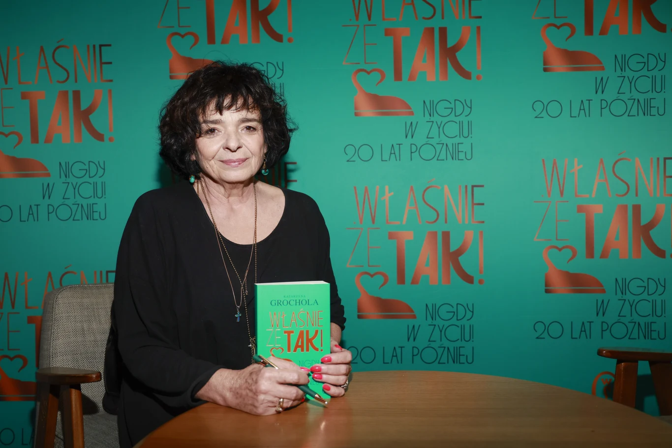 kobieta o ciemnych, kręconych włosach siedzi przy drewnianym stole, trzymając w dłoniach zieloną książkę z tytułem 'Właśnie Że Tak!', w tle turkusowa ścianka z tym samym tytułem oraz napisem 'Nigdy w życiu 20 lat później'