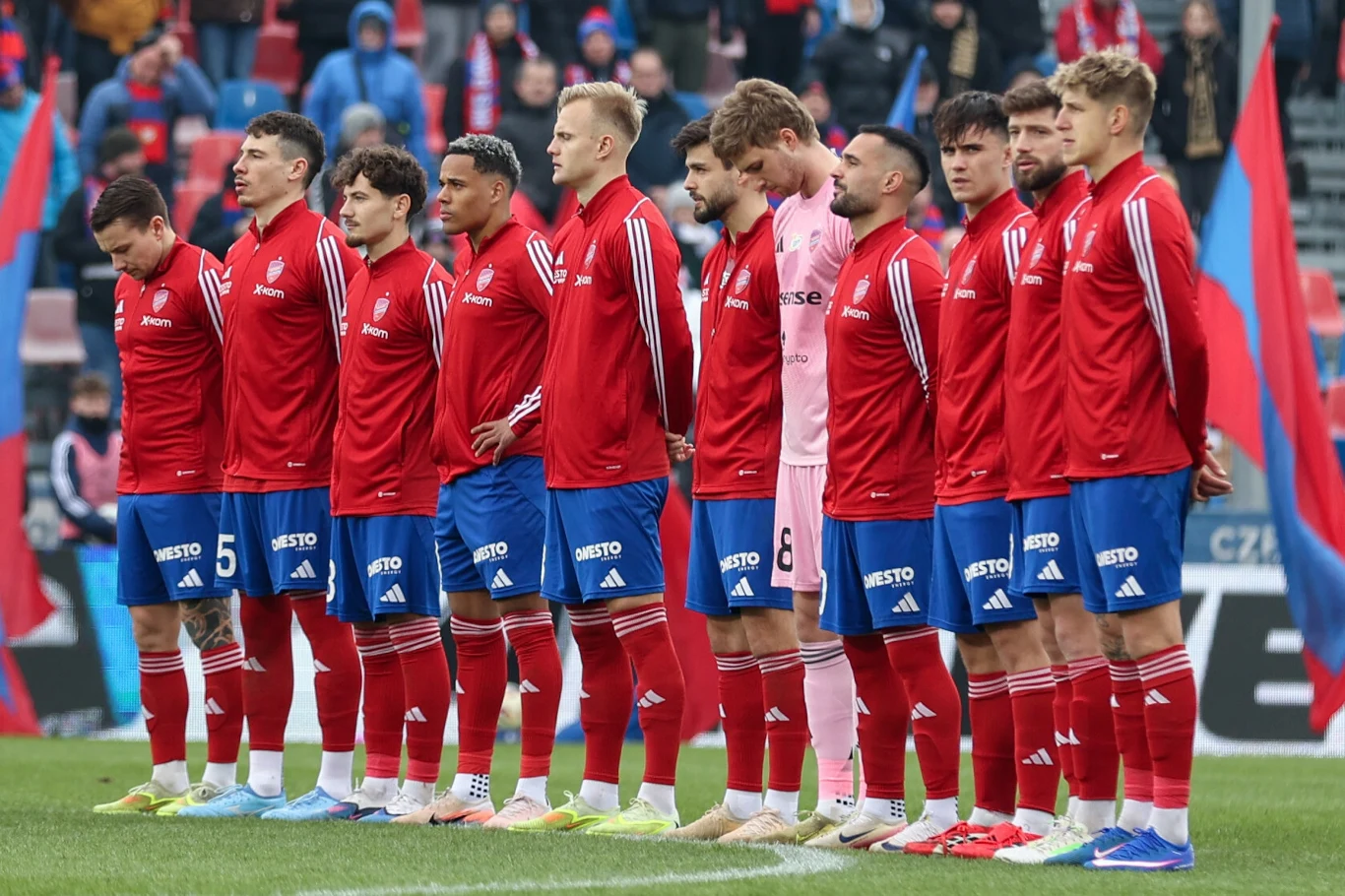 Piłkarze Rakowa Częstochowa jedenastu piłkarzy w czerwonych strojach sportowych oraz jeden zawodnik ubrany na różowo stoi w linii na stadionie, skupieni i poważni, w tle widoczni kibice i kolorowe flagi