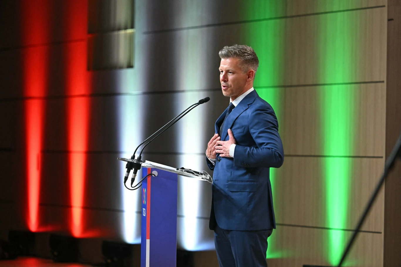Peter Magyar Peter Magyar w niebieskim garniturze przemawia przy mównicy, w tle ściana w barwach czerwonej, białej i zielonej