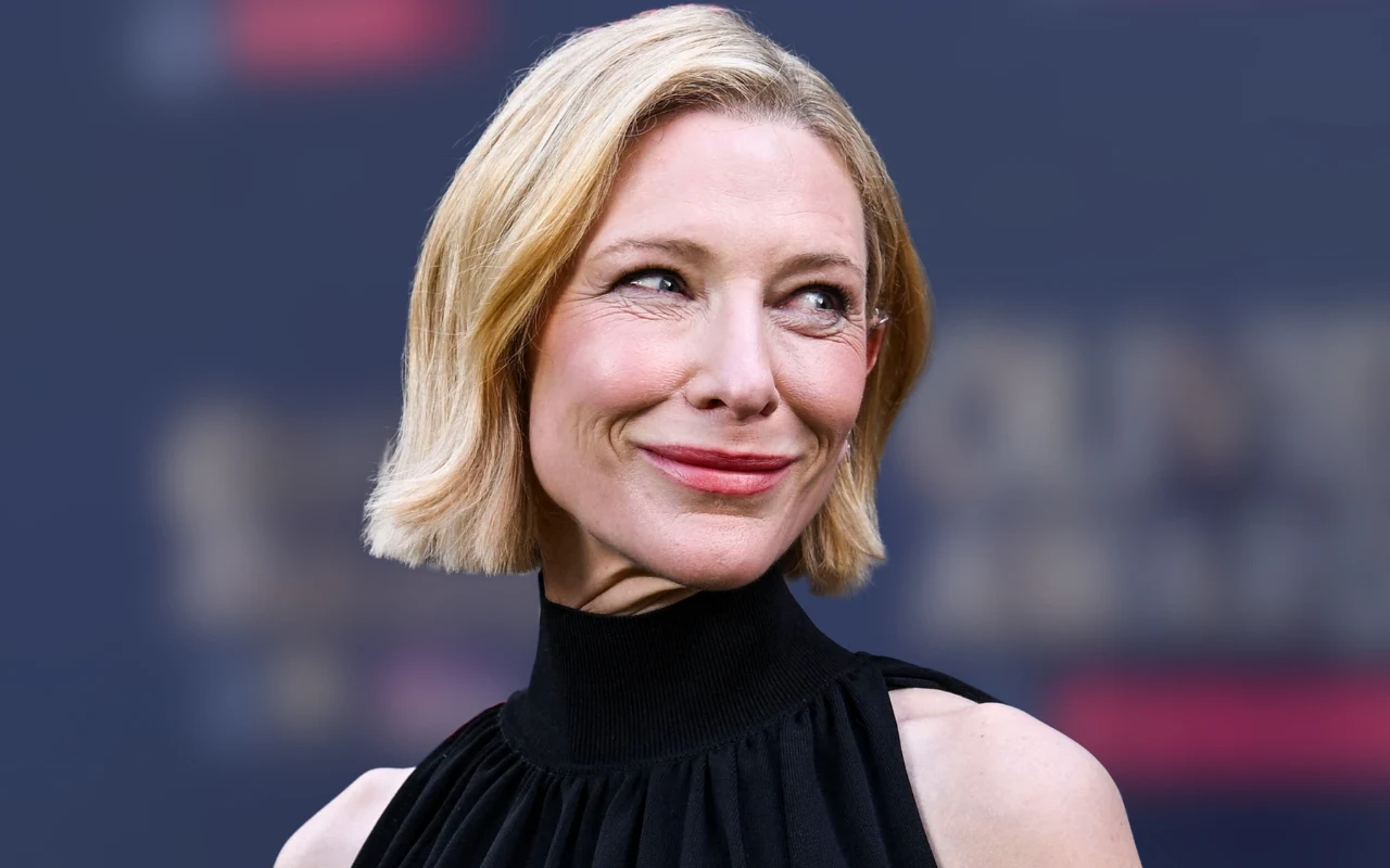 Kobieta o krótkich blond włosach uśmiecha się subtelnie, ubrana w elegancką czarną suknię z wysokim kołnierzem, na tle rozmytego ciemnego tła.