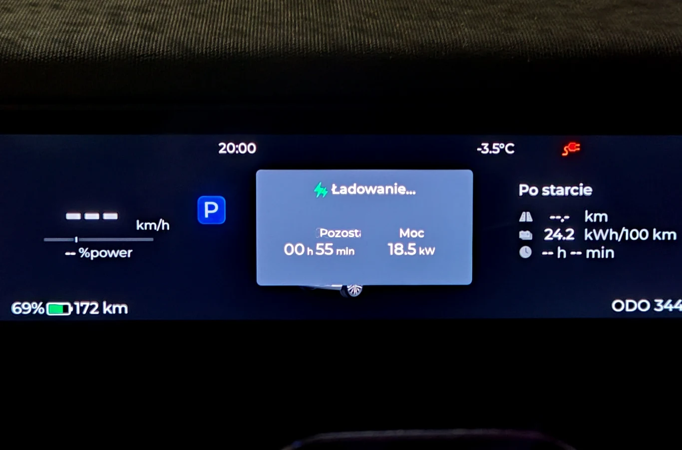 Cyfrowy wyświetlacz samochodu elektrycznego, środkowy panel informuje o trwającym ładowaniu baterii z pozostałym czasem 55 minut i mocą 18,5 kW, poziom naładowania 69% oraz zasięg 172 km, temperatura zewnętrzna -3,5°C, auto w trybie postoju (P), liczni...