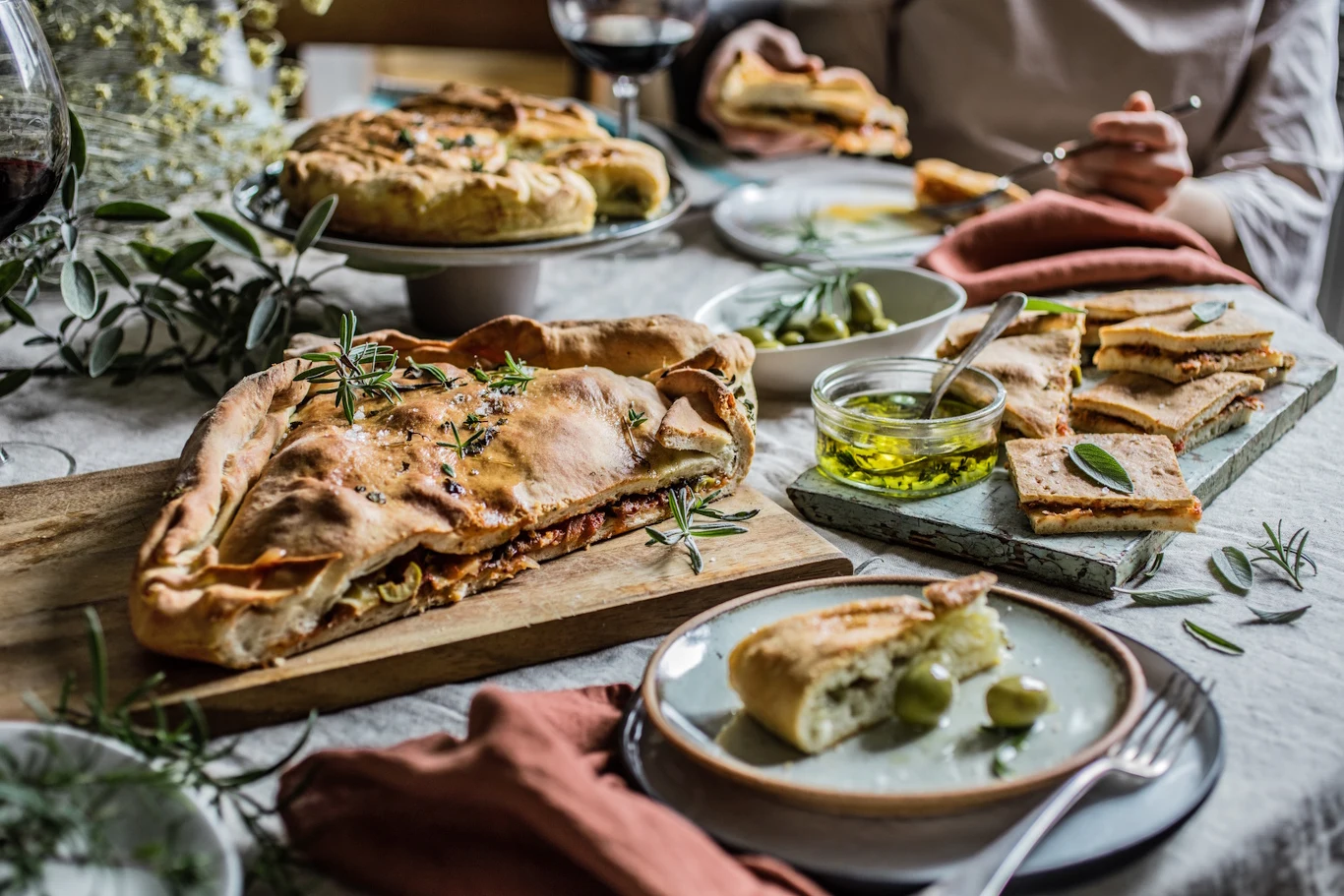 Ciasto kroi się i podaje jako przekąskę lub szybki lunch Stół zastawiony rustykalnym pieczywem na drewnianych deskach, focaccią z ziołami, kawałkami ciasta z oliwkami, miseczką oliwy z oliwek oraz gałązkami świeżego rozmarynu, wokół których siedzą osoby przygotowujące się do wspólnego posiłku.