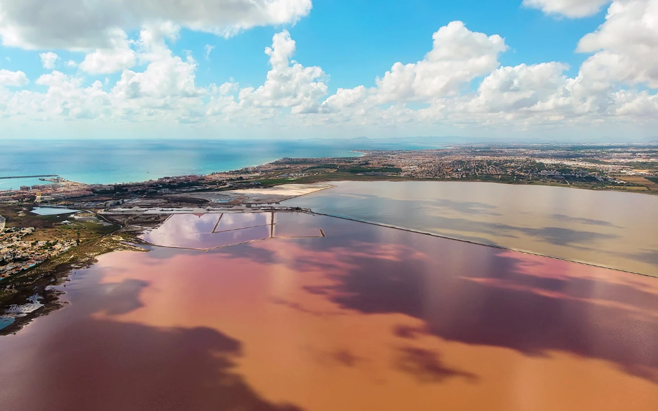Różowe i brązowe wody jeziora oddzielone groblą, odległa zabudowa miejska i błękitne niebo pokryte chmurami odbijającymi się w powierzchni jeziora.