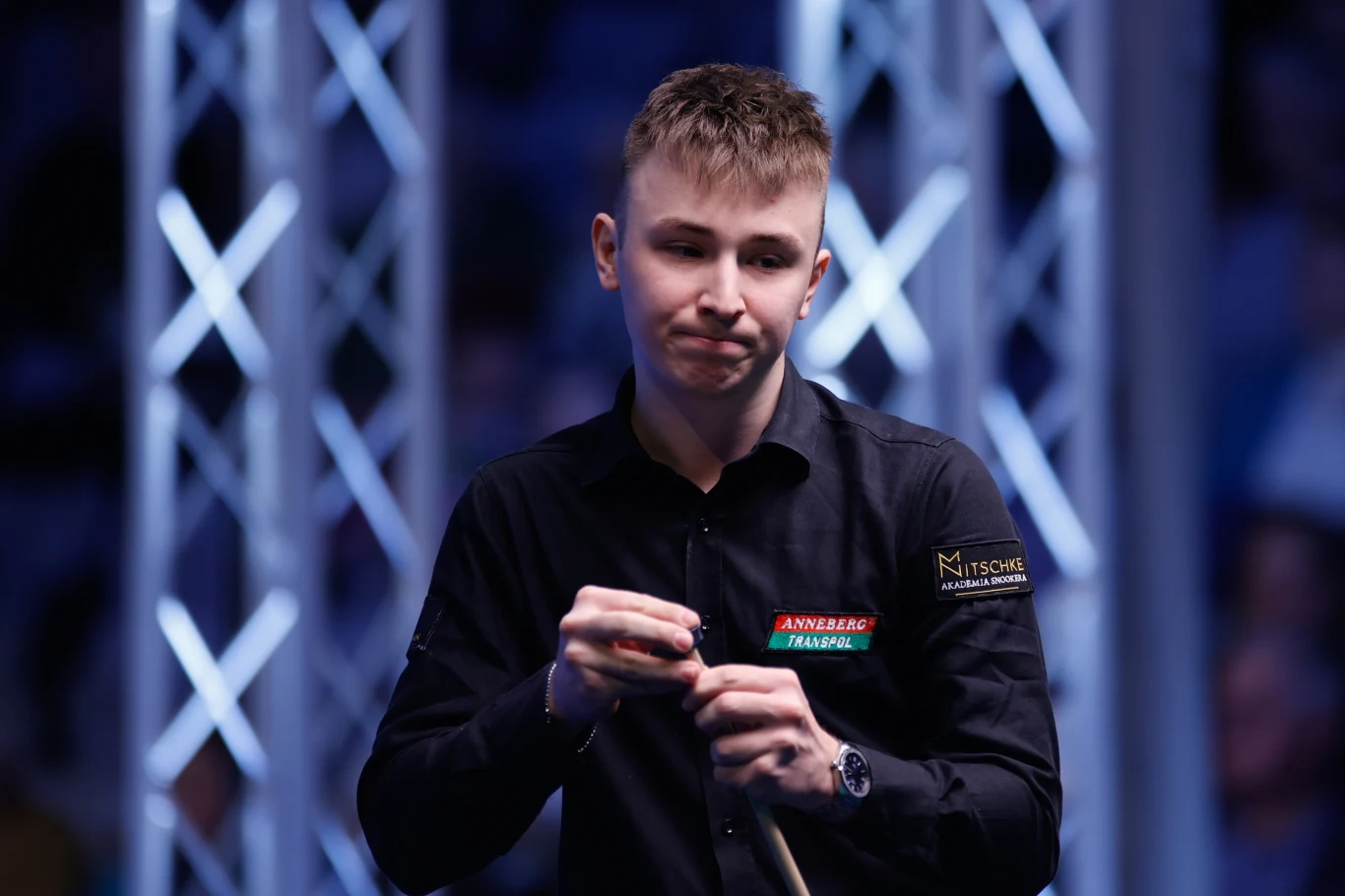 Młody mężczyzna w czarnej koszuli z naszywkami sponsorskimi trzyma kij bilardowy i kredę, a w tle widoczne są metalowe konstrukcje sceniczne oświetlone niebieskim światłem.