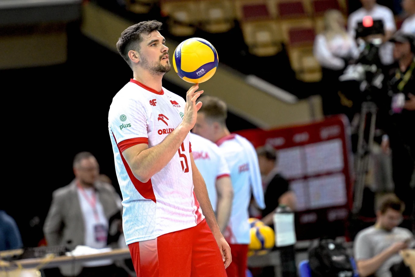 Mikołaj Sawicki siatkarz w biało-czerwonym stroju z logo Orlen balansuje piłkę na palcu, w tle inni zawodnicy i trybuny w hali sportowej