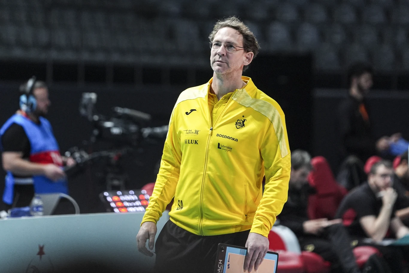 Stephane Antiga mężczyzna w żółtej sportowej kurtce z logo drużyny siatkarskiej stoi na hali sportowej, trzymając w ręku notatnik; w tle widoczne są osoby związane z obsługą wydarzenia oraz sprzęt do realizacji transmisji