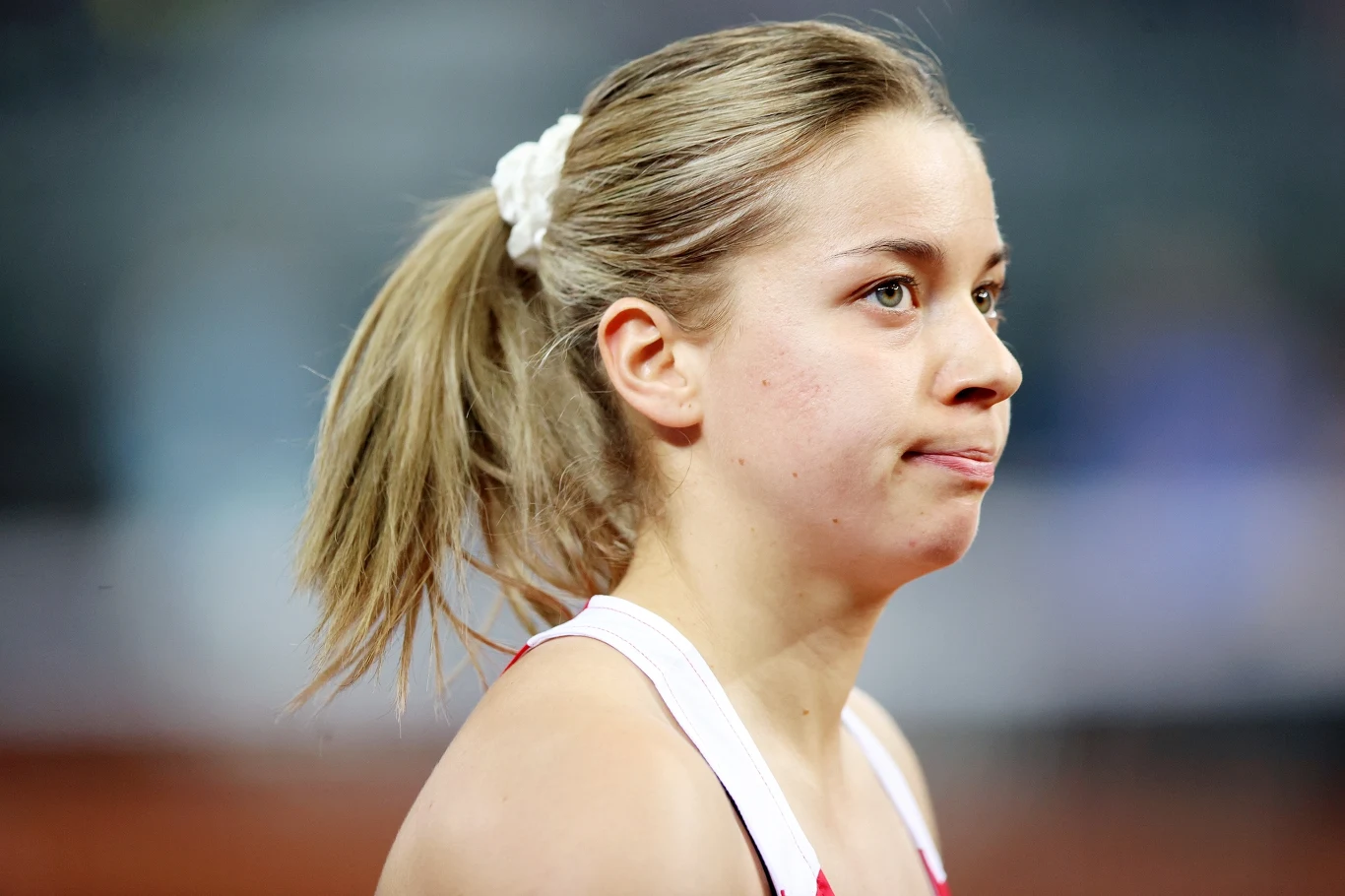 Młoda kobieta o jasnych włosach spiętych w kucyk białą gumką, ubrana w sportowy strój na tle rozmytego stadionu, z lekko skupionym wyrazem twarzy.