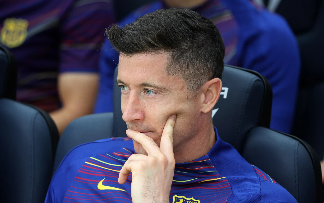 Mężczyzna w koszulce FC Barcelony siedzi na ławce rezerwowych, wspiera twarz na dłoni i patrzy zamyślony w bok.