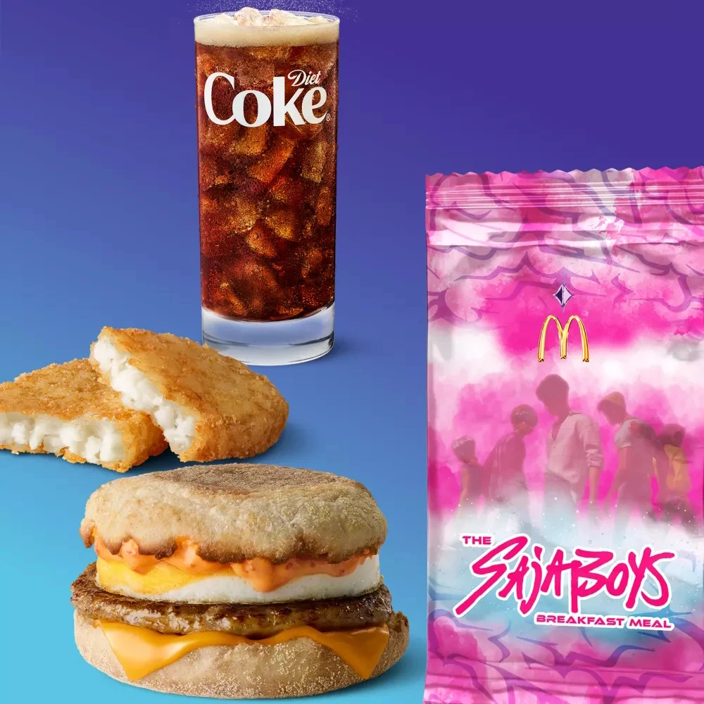 Zestaw śniadaniowy składający się z napoju Diet Coke w wysokiej szklance z lodem, dwóch chrupiących placków ziemniaczanych, kanapki śniadaniowej z jajkiem, serem i kiełbaską na angielskiej bułce oraz różowego opakowania promocyjnego McDonald's z napise...