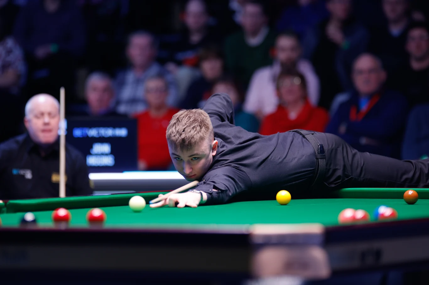 młody snookerzysta w czarnym stroju rozciąga się przy stole bilardowym, celując kijem w białą bilę, podczas gdy publiczność w tle obserwuje koncentrację gracza