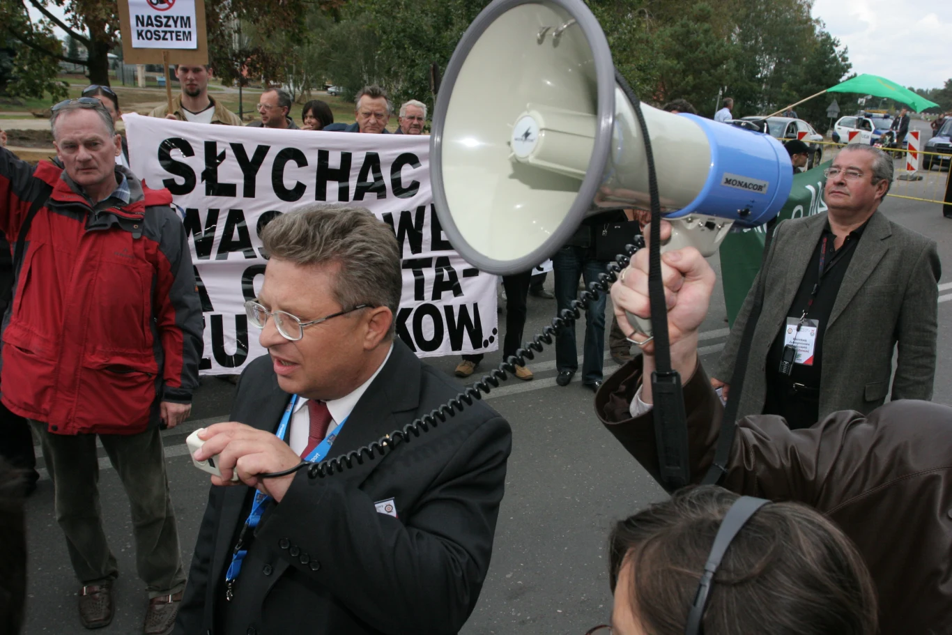 Grupa protestujących z transparentem zawierającym duże hasło, mężczyzna w garniturze przemawia do ludzi przez megafon, wokół stoją inni uczestnicy demonstracji obserwujący sytuację.