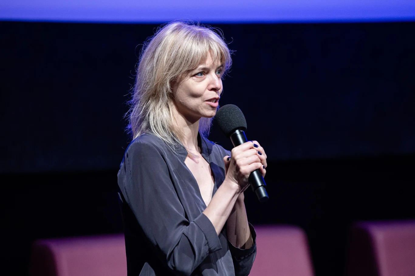 Joanna Łapińska, dyrektorka artystyczna Festiwalu Polskich Filmów Fabularnych w Gdyni Joanna Łapińska z mikrofonem na tle ciemnej sceny z fioletowym światłem i krzesłami w tle