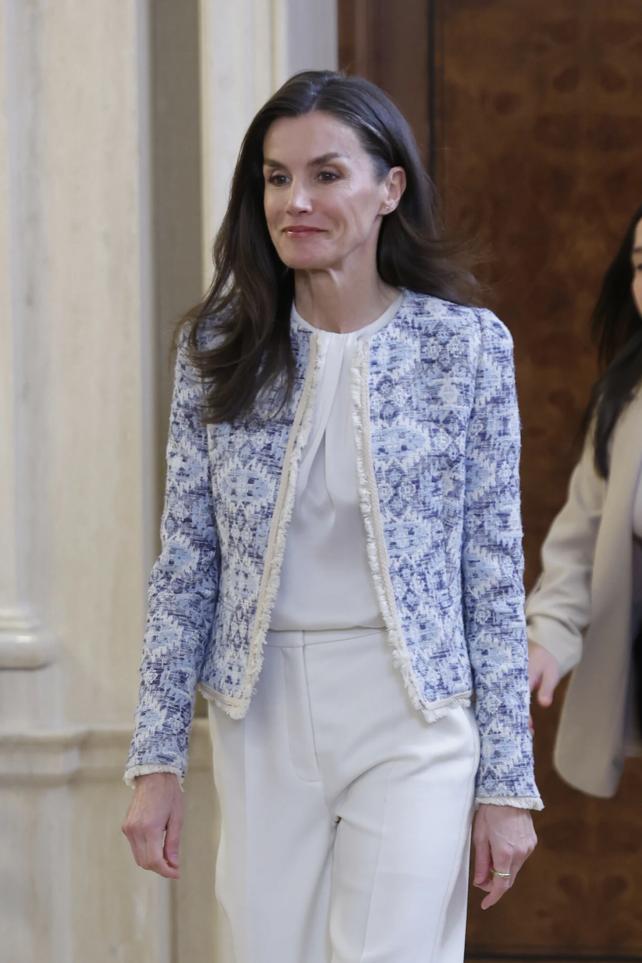 Królowa Letizia postawiła na dobrze znaną jej fanom kurtkę Kobieta o długich, ciemnych włosach ubrana w eleganckie, białe spodnie i białą bluzkę, na których nosi niebiesko-białą, wzorzystą marynarkę; w tle częściowo widoczna druga osoba w jasnym płaszczu.
