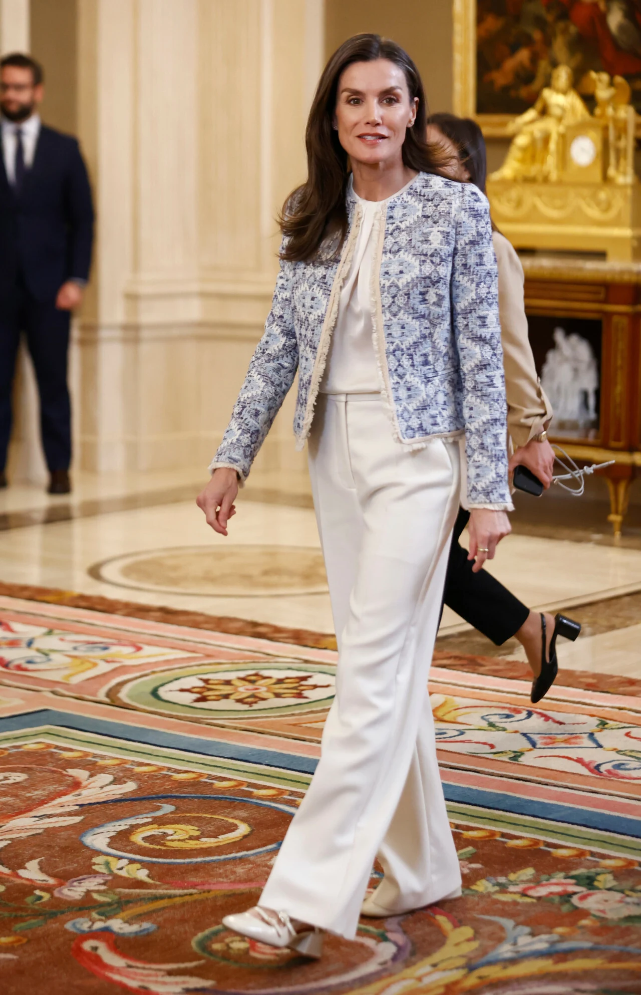 Stylizacja królowej doskonale sprawdzi się na wiosennych uroczystościach elegancka kobieta w białych szerokich spodniach i jasnej błękitno-białej marynarce, idąca po ozdobnym dywanie w reprezentacyjnym wnętrzu pałacowym z widocznym złotym zegarem w tle
