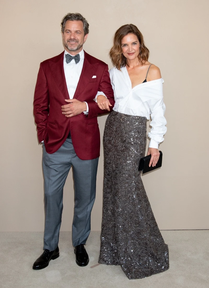 Elegancka para, mężczyzna w bordowej marynarce, białej koszuli i szarych spodniach z muchą, kobieta w białej rozpiętej koszuli z widocznym ramiączkiem i długiej, błyszczącej spódnicy, trzymająca czarną kopertówkę, oboje uśmiechnięci, pozujący na tle ja...