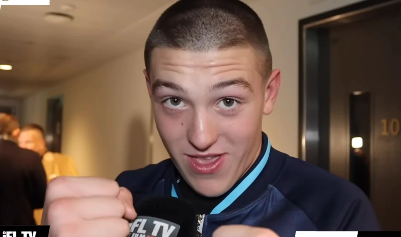 uśmiechnięty młody mężczyzna w sportowej kurtce trzyma zaciśnięte pięści w geście bokserskim, na pierwszym planie widoczny mikrofon z logo, w tle korytarz i rozmyte postacie