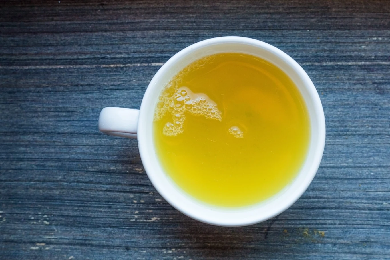 biała filiżanka wypełniona jasnożółtym naparem, ustawiona na ciemnym drewnianym blacie