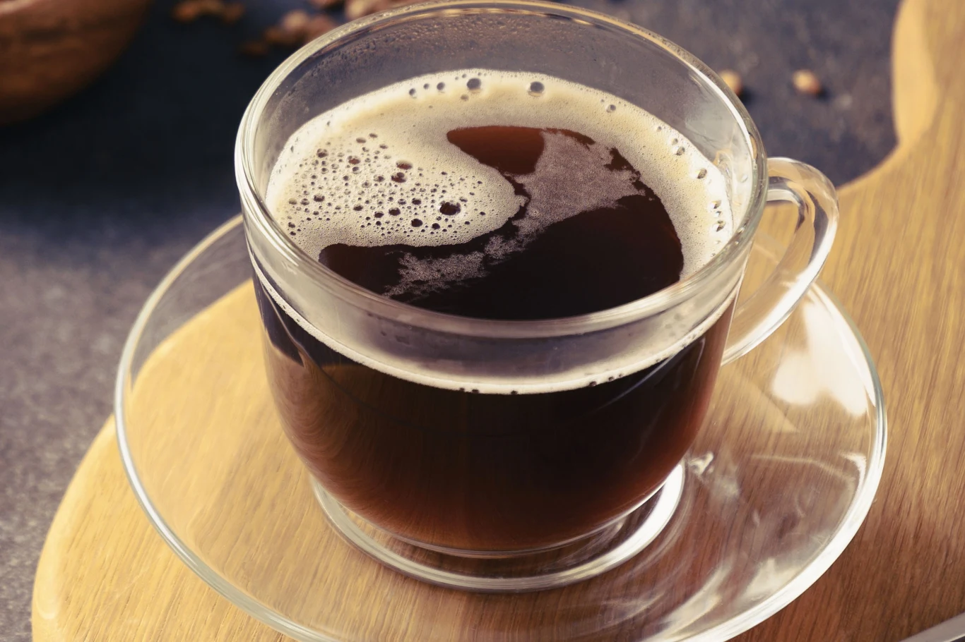 Kawa jęczmienna wspaniale wspiera jelita Szklanka kawy jęczmiennej z pianką na przezroczystym spodku, ustawiona na drewnianej desce.