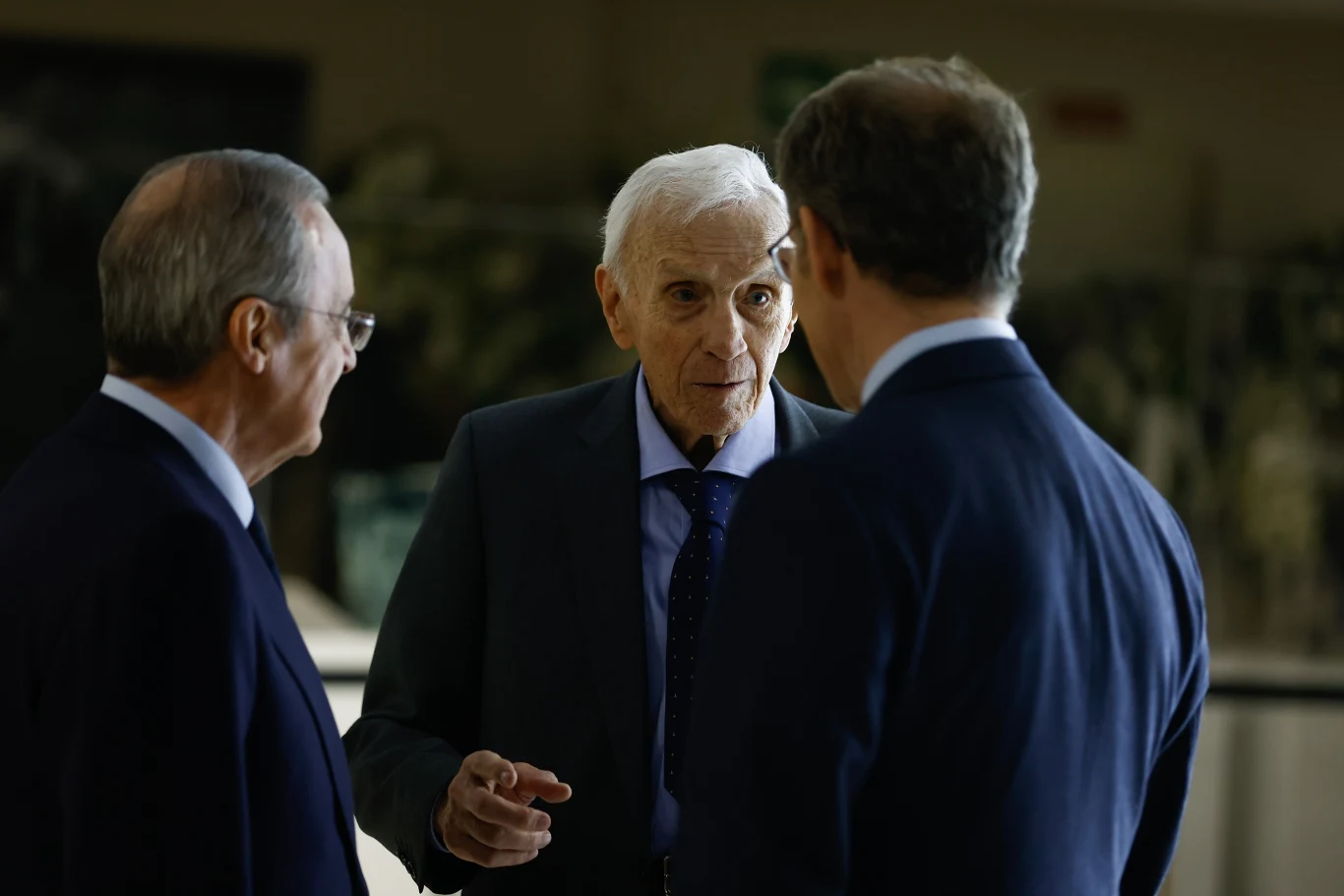 Jose Emilio Santamaria Trzech mężczyzn w garniturach prowadzi poważną rozmowę, jeden z nich gestykuluje dłonią, skupiając uwagę pozostałych rozmówców.