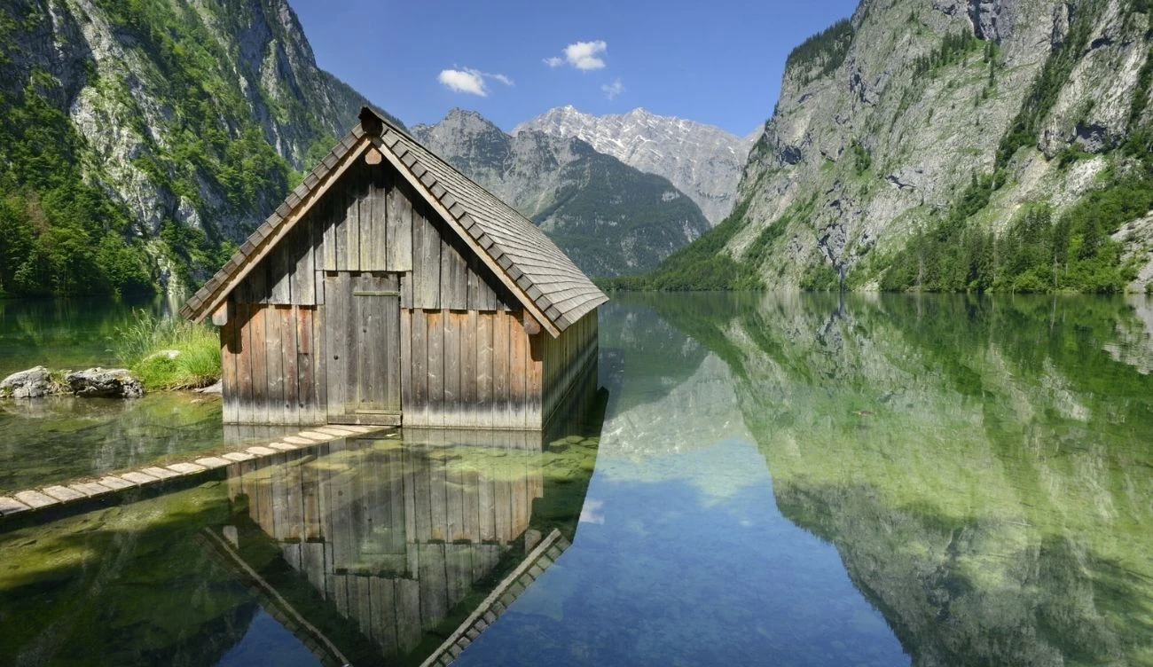 Obersee to jedno z najpiękniejszych jezior w Niemczech Drewniana chatka przy jeziorze Obersee częściowo zanurzona w wodzie, otoczona górami i odbiciem w tafli jeziora.