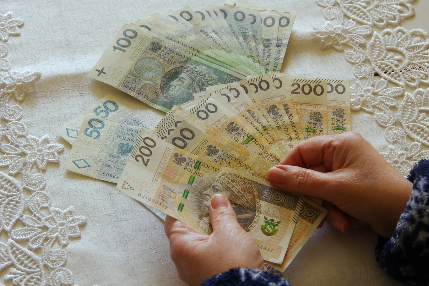 Jak oszczędzać na emeryturę? Osoba trzyma wachlarz polskich banknotów na białym, koronkowym obrusie.