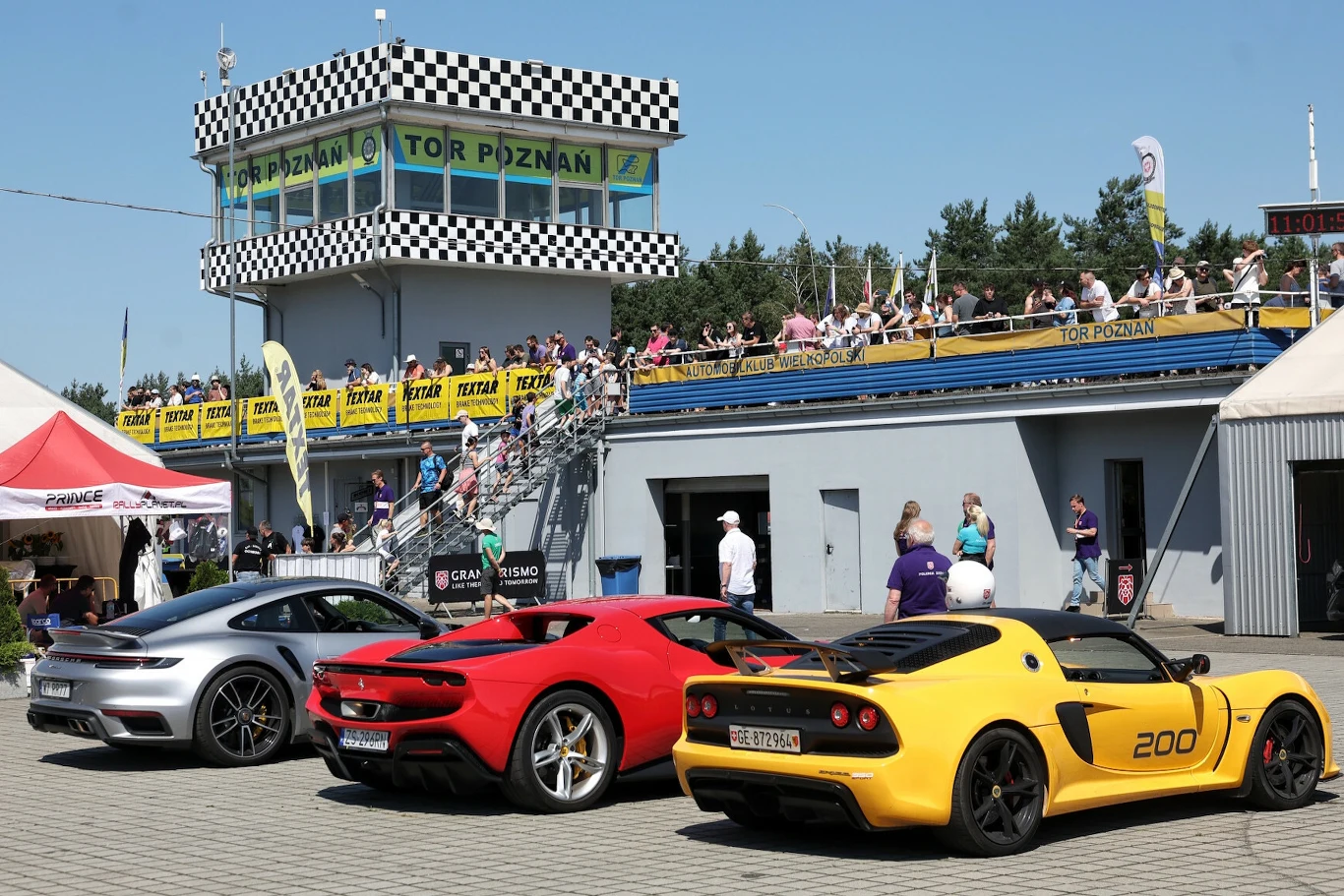 trzy sportowe samochody w jaskrawych kolorach stoją zaparkowane przed budynkiem toru wyścigowego z charakterystyczną szachownicą i trybuną wypełnioną kibicami, żółtymi banerami oraz napisem 'Tor Poznań', w tle osoby spacerujące i obserwujące wydarzenie