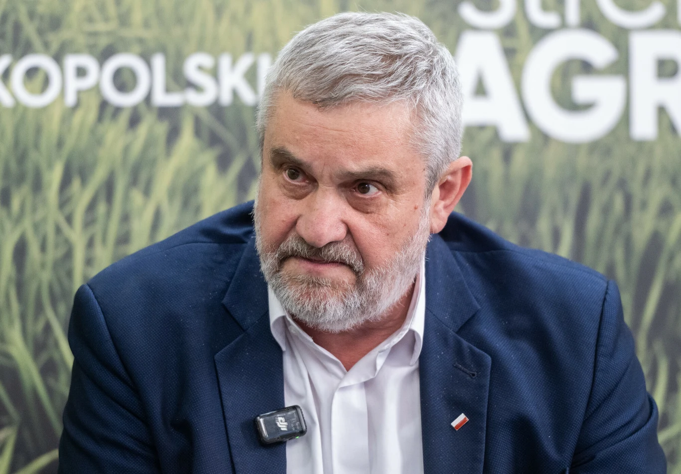 Jan Krzysztof Ardanowski, były minister rolnictwa i rozwoju wsi Jan Krzysztof Ardanowski w białej koszuli i granatowej marynarce z flagą Polski, z mikrofonem, na tle grafiki pola.