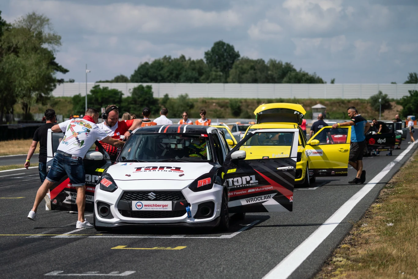 kilka sportowych samochodów ustawionych na torze wyścigowym, wokół których kręcą się członkowie zespołów, przygotowując auta do startu; pierwszy plan zajmuje biały samochód z otwartymi drzwiami i widocznymi sponsorami, w tle kolejne auta w jaskrawych k...