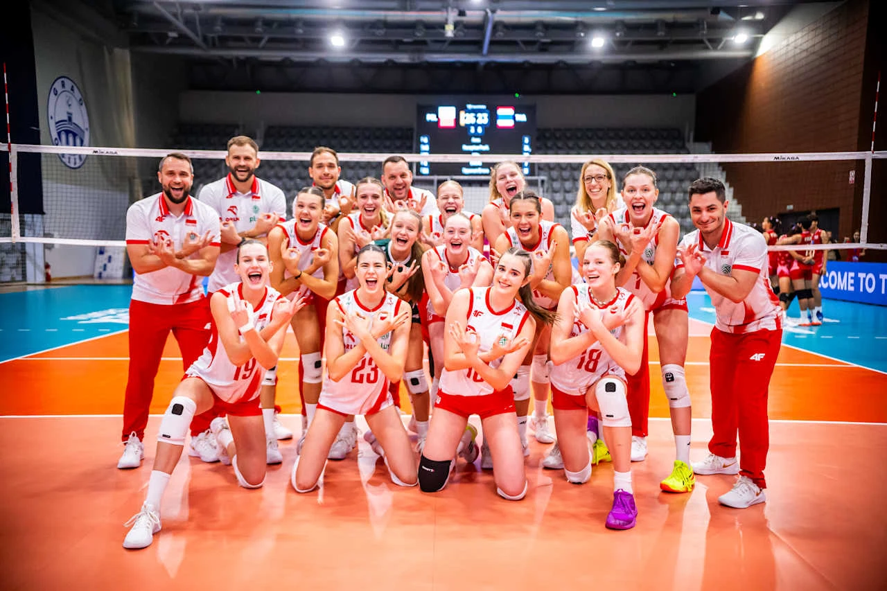 drużyna siatkarska kobiet w biało-czerwonych strojach pozuje na środku boiska, wszyscy zawodnicy i sztab szkoleniowy uśmiechają się i wykonują gest zwycięstwa, atmosfera pełna radości i sportowej energii