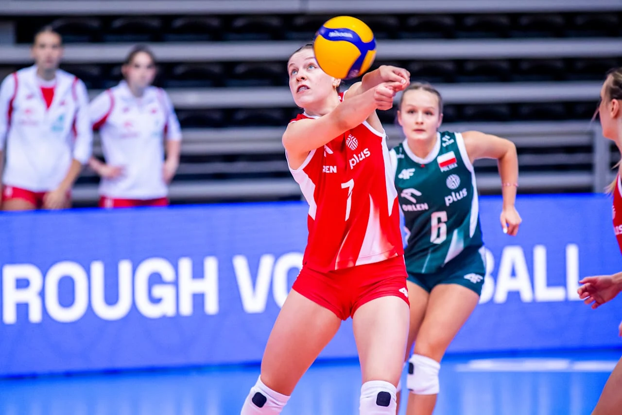 siatkarka w czerwono-białym stroju odbija piłkę sposobem dolnym, obok zawodniczki z przeciwnej drużyny w zielonym stroju oraz zawodniczki w tle przy siatce