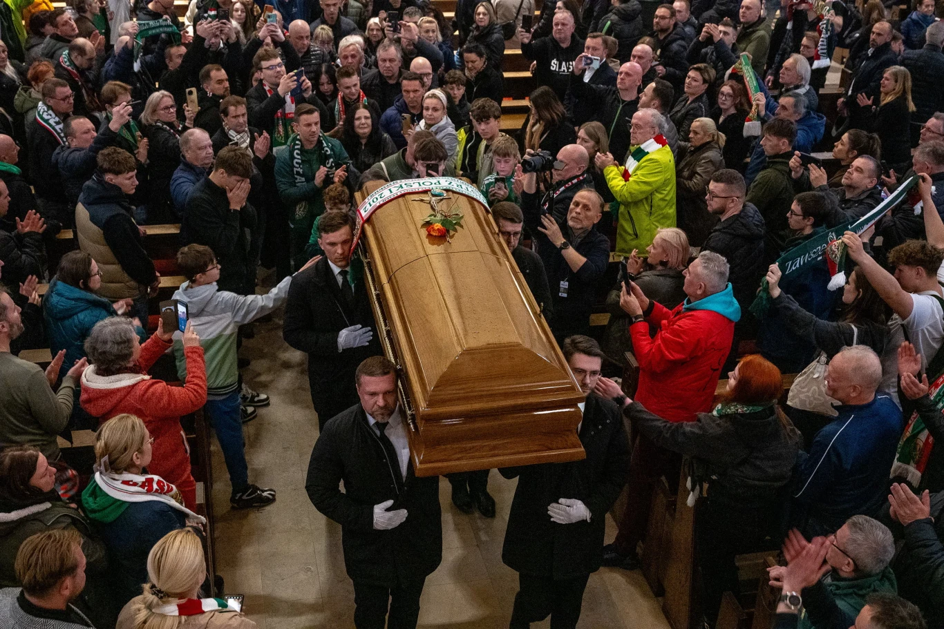 trumna niesiona przez sześciu mężczyzn wśród tłumu ludzi w kościele, wokół żałobnicy w różnym wieku, wielu z nich ubranych w barwy klubowe i szaliki, niektórzy biją brawo lub rejestrują moment telefonem, atmosfera skupienia i pożegnania