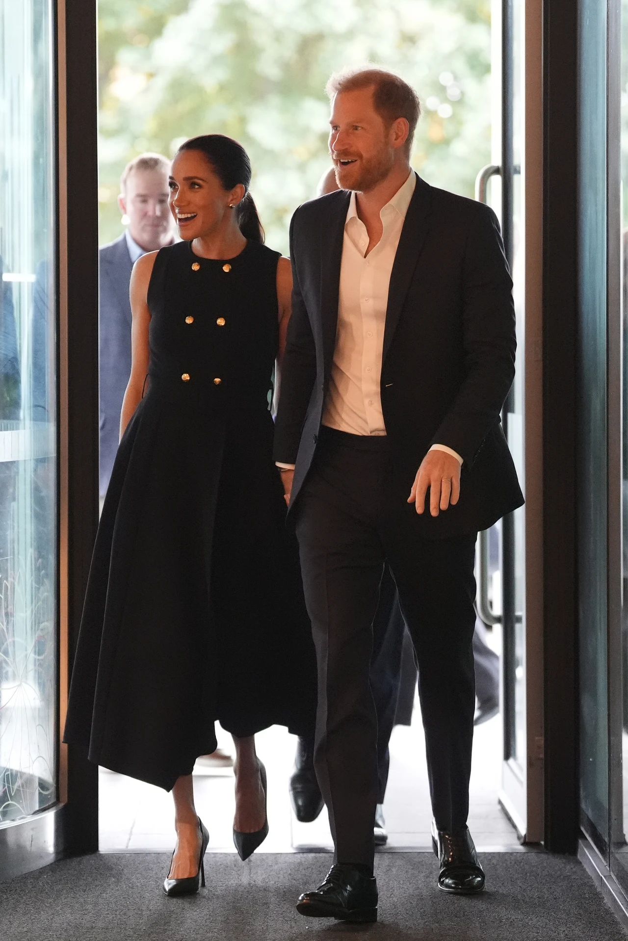 Para elegancko ubranych osób wchodzi do nowoczesnego budynku, trzymając się za ręce; kobieta ma na sobie czarną suknię midi z podwójnym rzędem złotych guzików, a mężczyzna garnitur i białą koszulę bez krawata.