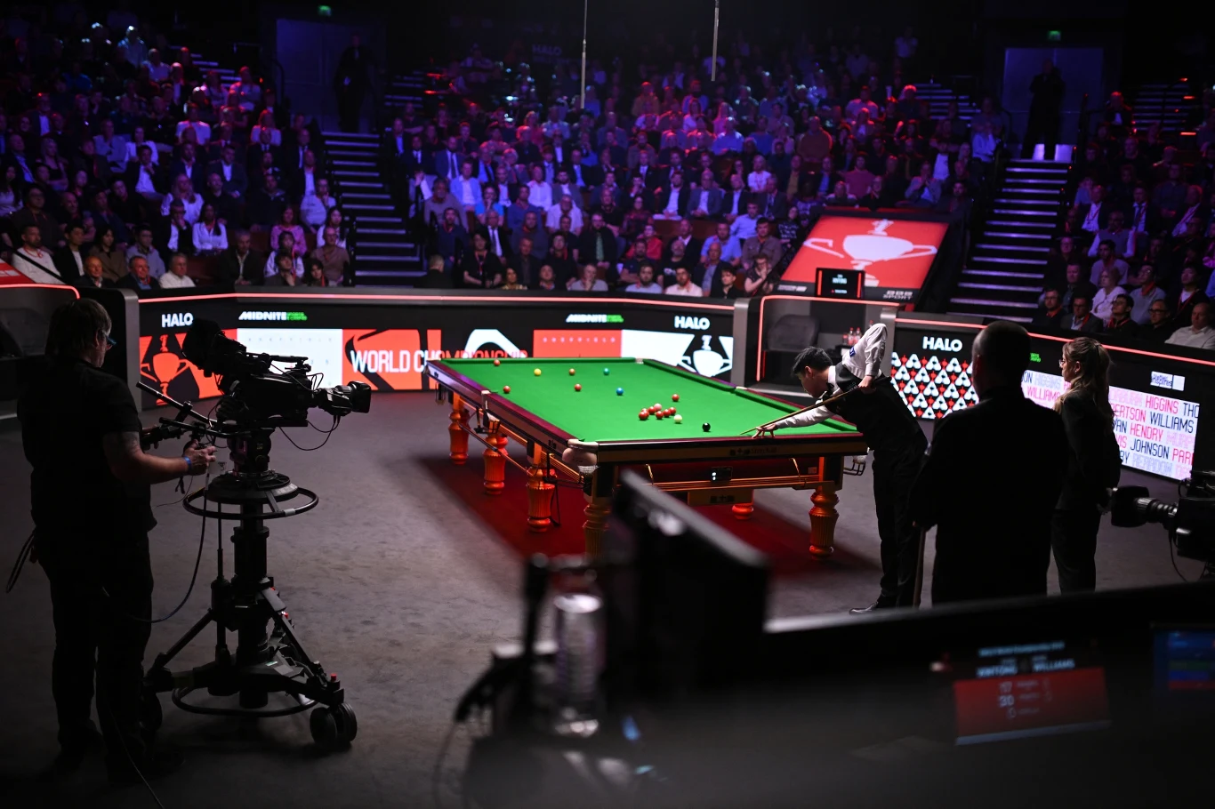 Crucible Theatre - scena decydujących zmagań o mistrzostwo świata w snookerze zielony stół bilardowy ustawiony w centrum oświetlonej areny, wokół którego kilku zawodników i kamerzysta przygotowują się do nagrania, a na trybunach zgromadzona jest licznie publiczność