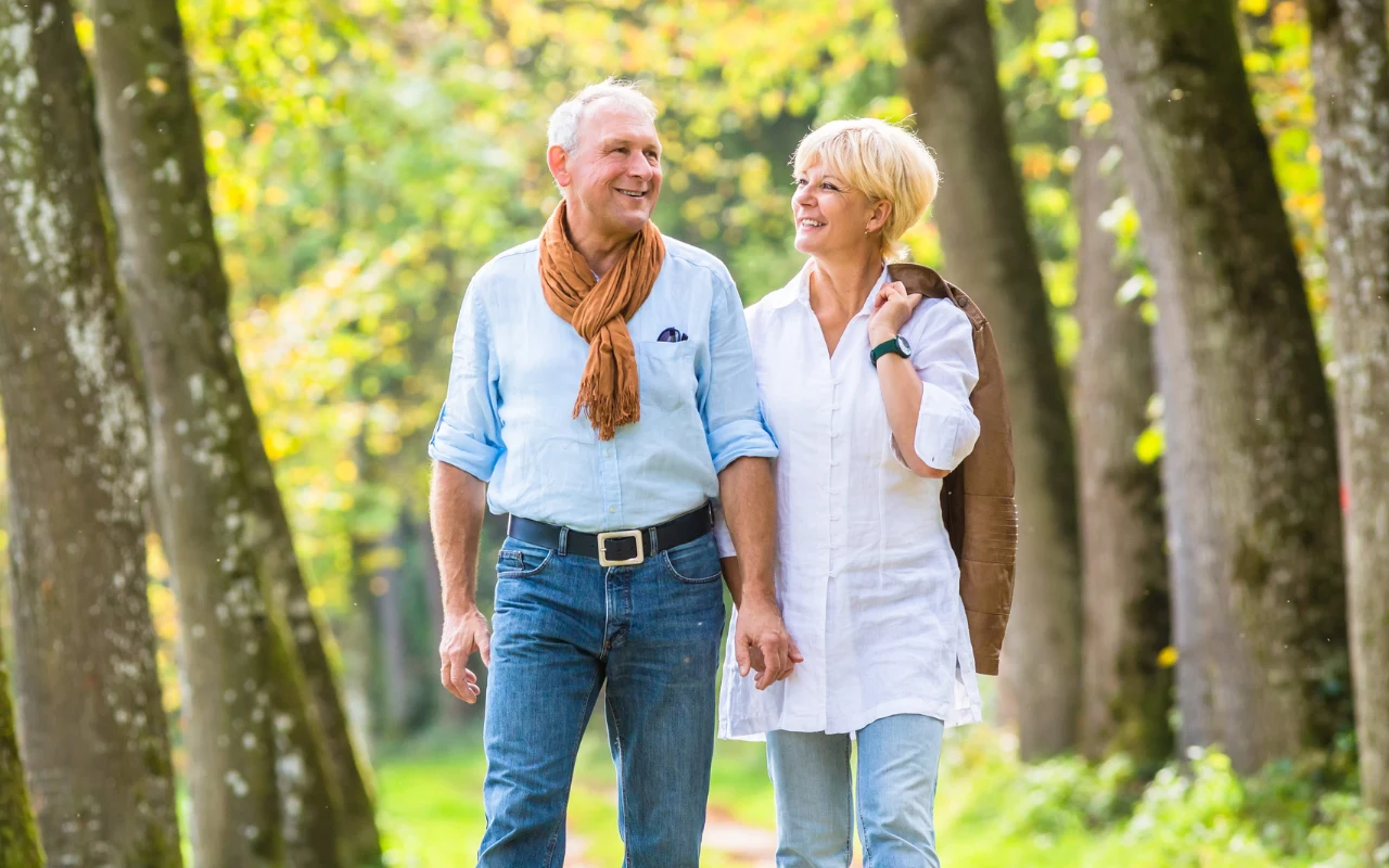 uśmiechnięta para starszych osób trzyma się za ręce, spacerując razem alejką w parku otoczoną drzewami w słoneczny dzień