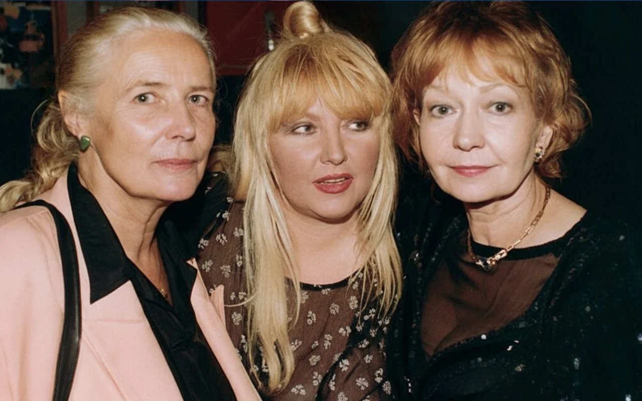 Trzy kobiety stoją blisko siebie, patrząc w różne strony. Środkowa postać ma długie blond włosy i jasny makijaż, a pozostałe dwie mają jasne włosy spięte do tyłu oraz noszą eleganckie, ciemne ubrania.