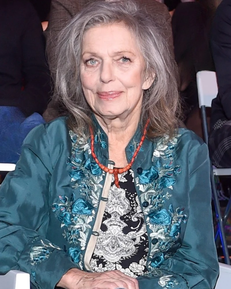 starsza kobieta z długimi, siwymi włosami siedzi na krześle, ubrana w elegancką, turkusową marynarkę z kwiatowym haftem i czarną bluzką w jasny wzór, na szyi ma czerwony naszyjnik