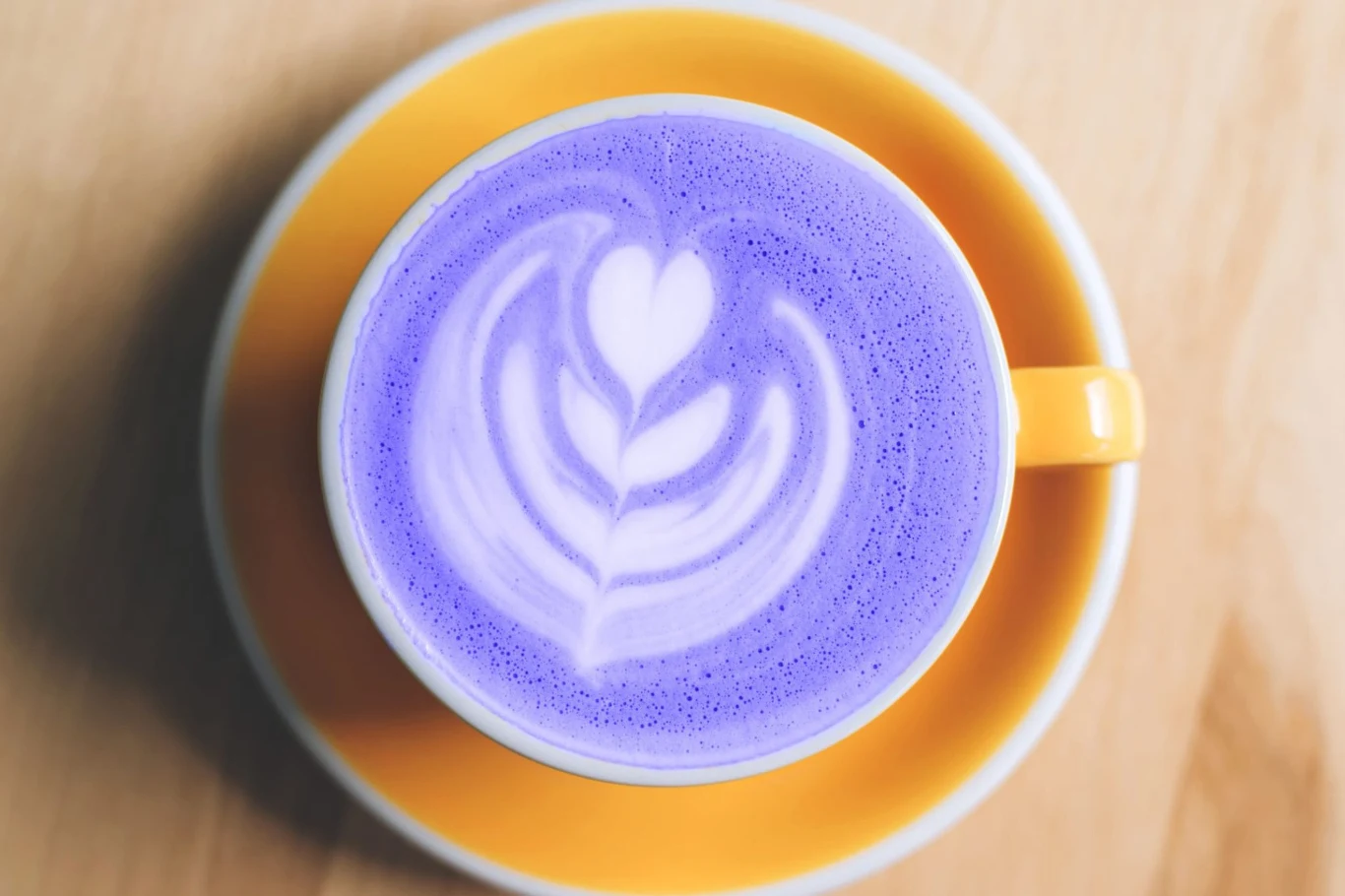 Fioletowa latte zachwyca smakiem i wyglądem Filiżanka z żółtym uchem wypełniona fioletowym ube latte z latte art w kształcie liścia i serc na piance.