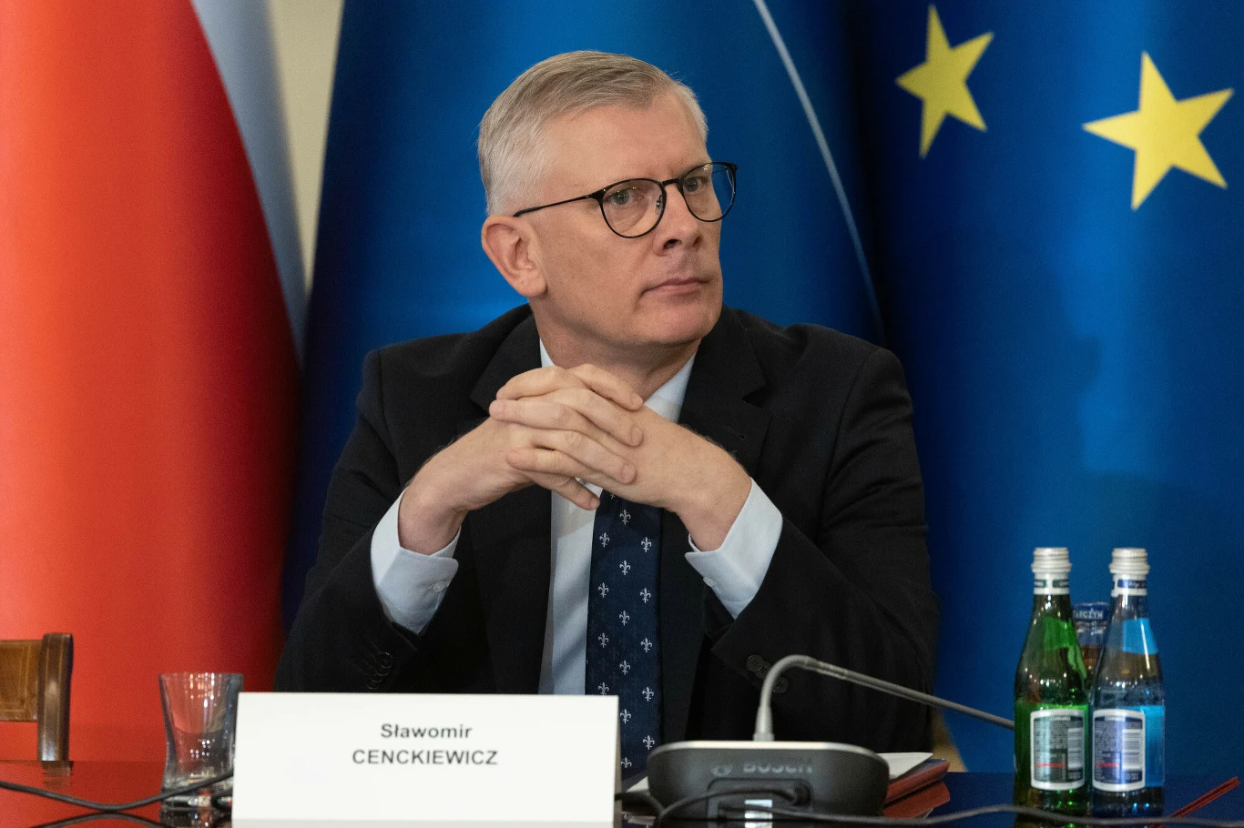 Były już szef Biura Bezpieczeństwa Narodowego prof. Sławomir Cenckiewicz Mężczyzna w okularach siedzi przy stole konferencyjnym, ubrany w garnitur i krawat, z dłońmi złożonymi przed sobą; w tle widoczne flagi Polski i Unii Europejskiej oraz tabliczka z nazwiskiem 'Sławomir Cenckiewicz' ustawiona przed nim.