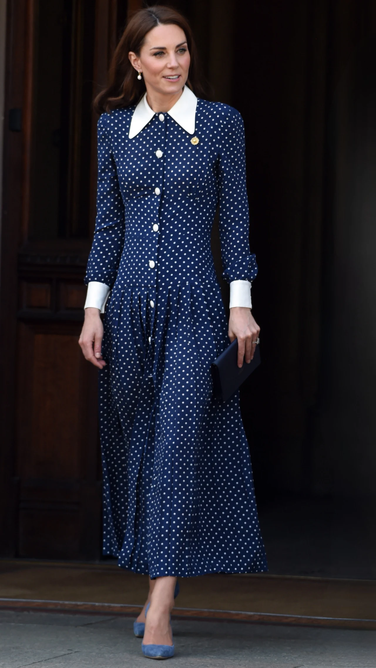 kobieta w granatowej sukience w białe kropki z białym kołnierzykiem i mankietami, trzyma granatową kopertówkę i ma na nogach niebieskie buty na obcasie