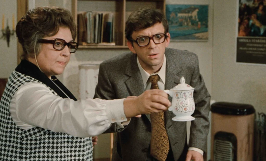 Kadr z filmu "Jak utopić doktora Mraczka?", fot. Timeless Film Festival Warsaw Kobieta w okularach podaje filiżankę mężczyźnie w garniturze i okularach, oboje mają poważne miny, w tle półka z książkami i plakat na ścianie.