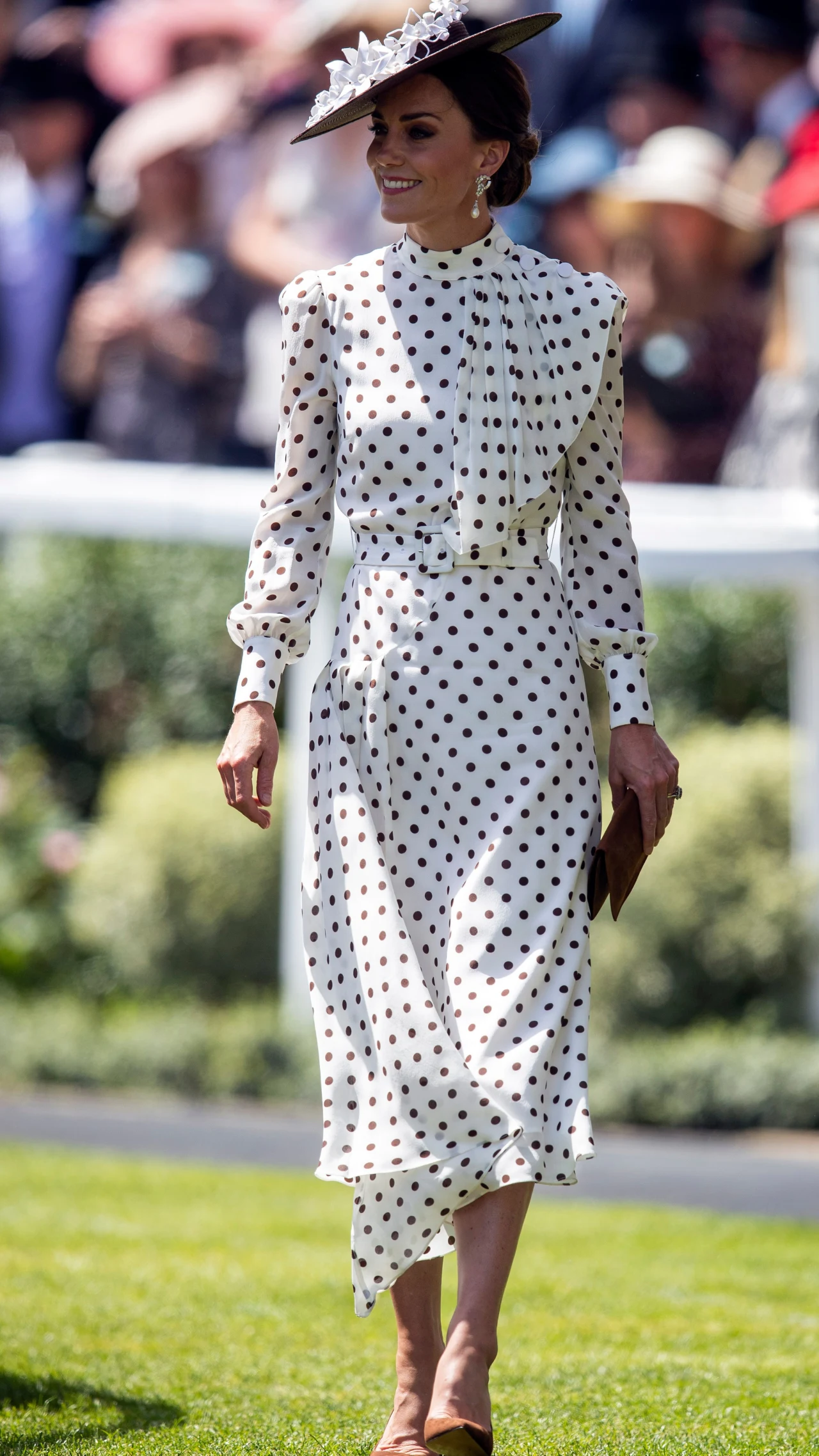 kobieta w eleganckiej białej sukience w czarne grochy, ozdobiona efektownym kapeluszem z piórami, trzymająca kopertówkę, spaceruje po trawniku podczas formalnego wydarzenia