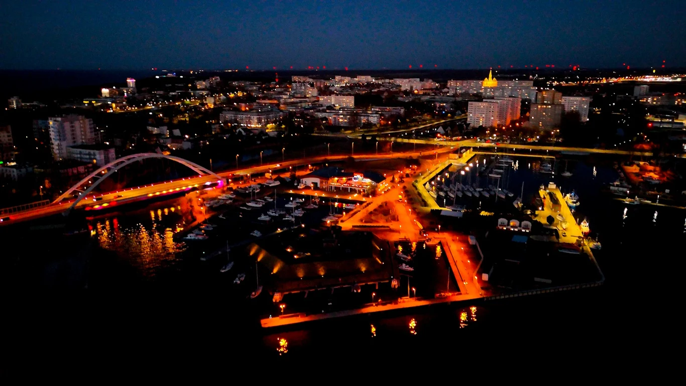 Nocne światła miasta tworzą malowniczy widok nad Bałtykiem most oświetlony pomarańczowym światłem prowadzący nad mariną z licznymi jachtami, w tle rozświetlone budynki miejskie i jasno podświetlony gmach z żółtym dachem stanowiący dominantę krajobrazu nocnego miasta