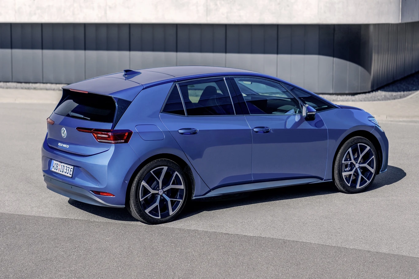 niebieski samochód elektryczny Volkswagen ID.3 zaparkowany na tle nowoczesnego, betonowego budynku, widok z tyłu i boku podkreśla aerodynamiczną sylwetkę oraz charakterystyczne lampy LED
